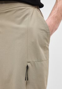 Gros plan sur un pantalon beige avec une main partiellement dans la poche et une petite tirette noire suspendue à une couture.