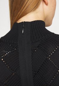Sort strikket sweater med høj krave, der har et struktureret mønster og en lynlås i ryggen. Strikken har et let åbent væv.