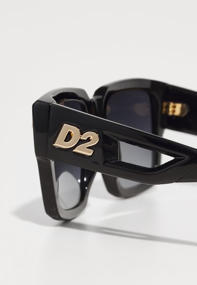 Gli occhiali da sole neri presentano un design massiccio con un logo "D2" dorato sulla montatura. Le lenti sono scure e rettangolari, con una finitura lucida.
