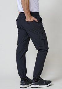 Moški nosi črne joggers hlače s elastičnimi manšetami, črne superge in belo majico, stoji z eno roko v žepu.