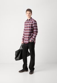 Camicia a quadri nei toni del rosso e navy, abbinata a pantaloni in velluto a coste neri e sneaker nere. Il modello tiene una borsa a tracolla nera.