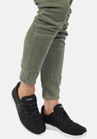 Schwarze Filzsneaker mit weißen Gummisohlen, ausgestattet mit Schnürverschluss und einem Logo-Akzent an der Seite, kombiniert mit olivgrünen Hosen.