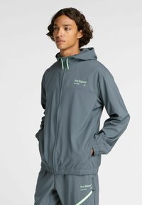 Joven con cabello rizado que lleva una chaqueta con capucha gris de New Balance y pantalones a juego, con las manos en los bolsillos, mirando hacia la izquierda.