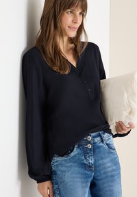 Blouse noire avec un décolleté en V et des boutons, manches longues et poignets froncés, associée à un jean bleu clair. Le modèle tient un coussin matelassé de couleur crème.