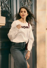 Witte sweatshirt met bordeauxrode "mom" tekst, gecombineerd met een donkerdenim rok. Model poseert tegen een versierde deur met decoratieve details.