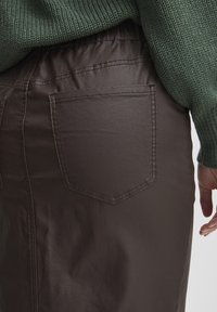 Jupe marron à taille élastique avec une texture lisse, dotée de deux poches arrière avec des accents cousus. Le tissu semble léger et décontracté.