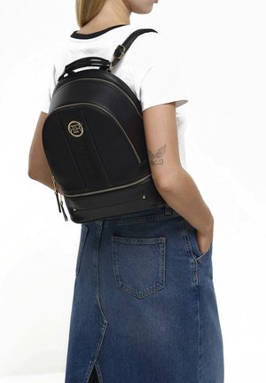 Donna con maglietta bianca e gonna in denim con uno zaino nero in pelle con cerniere dorate e logo sulla schiena.