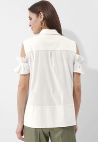 Witte blouse met korte, gerimpelde mouwen en een geknoopte kraag. Bevat een rugplooi en een iets langere zoom, gecombineerd met olijfgroene broeken.