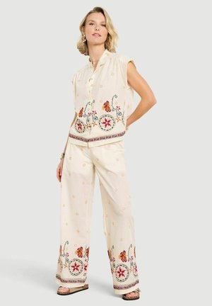 Kobieta w kremowej bluzce bez rękawów i szerokich spodniach z czerwonym kwiatowym haftem, stoi z jedną ręką na biodrze.