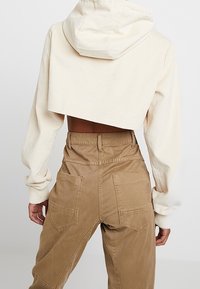 Osoba v béžové zkrácené kordové mikině a hnědých kalhotách s vysokým pasem a velkými zadními kapsami, viděná zezadu.
