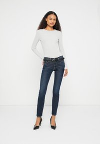 Jean skinny en denim bleu foncé avec une finition texturée, associé à un haut ajusté à manches longues gris clair et des talons pointus noirs.