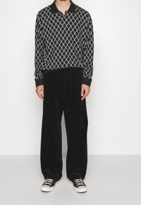 Polo noir à manches longues avec un motif ondulé blanc, associé à un pantalon large en velours côtelé noir et des baskets noires avec des semelles blanches.