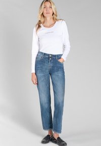 Haut à manches longues côtelé blanc avec texte, jeans en denim bleu taille haute, jambe droite, accentués de boutons dorés et de détails délavés, chaussures noires à enfiler.