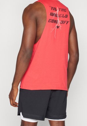 Mann trägt korallenrotes ärmelloses Shirt mit schwarzem Text ‚TIL THE WHEELS COME OFF‘ und schwarze Shorts vor schlichtem hellem Hintergrund.