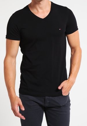 Homme portant un T-shirt noir ajusté à col en V avec un petit logo rouge, blanc et bleu sur la poitrine, associé à un pantalon foncé, une main dans la poche.