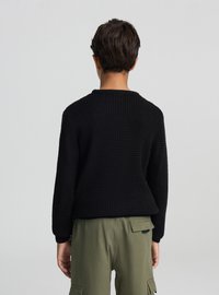 Persona con capelli castani corti che indossa un maglione nero a trama e pantaloni cargo verde oliva, vista di schiena su uno sfondo grigio chiaro uniforme.