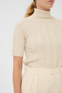 Kvinde iført en beige ribbet kortærmet turtleneck sweater, der er stoppet ned i højtaljede cremefarvede plissert bukser.