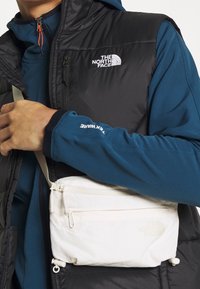 Czarna kamizelka na niebieskiej bluzie z kapturem; ręka trzyma kremową torebkę na ramię z zamkiem i logo; miękka tkanina i minimalistyczny design.