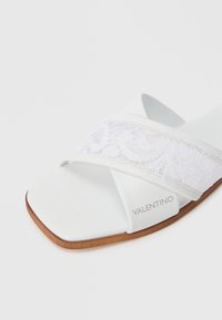 Hvit sandal med kryssdesign og snøredetaljer, glatt skinnøvre, og flat brun såle. Merkevarelogo synlig på siden.