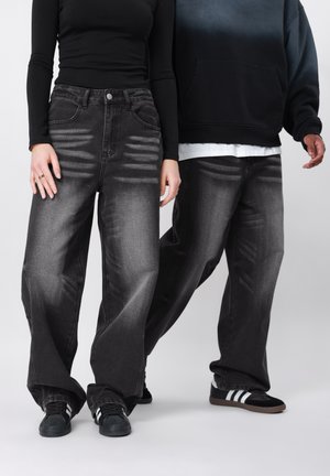 Deux personnes portant des jeans noirs délavés à jambes larges et des baskets noires, debout devant un fond gris clair.