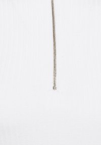 Witte top met een ritssluiting aan de voorkant, een geribbelde textuur en een rechte silhouet. De ritssluiting is van metaal met een zilverkleurige afwerking.