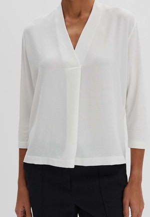 Femme portant un chemisier blanc ample à col en V avec manches 3/4 et un pantalon noir sur un fond uni.