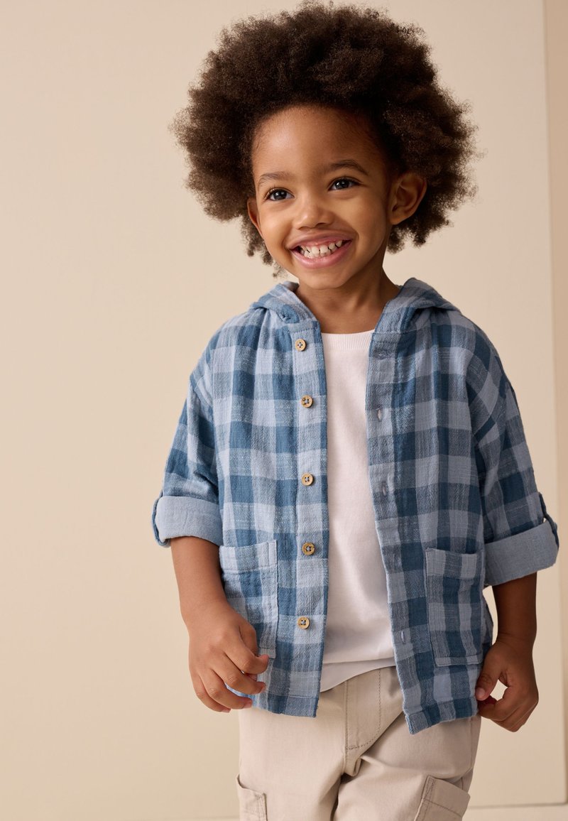 Enfant souriant aux cheveux bouclés portant une chemise à carreaux bleue boutonnée sur un t-shirt blanc et un pantalon beige, debout devant un fond uni.