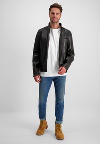 Schwarze Lederjacke mit hohem Kragen, getragen über einem weißen T-Shirt, kombiniert mit blauen Jeans und braunen Schnürstiefeln. Einfache Hintergrund.