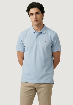 Hombre con camisa polo de manga corta azul claro y pantalones beige, de pie con las manos detrás de la espalda sobre un fondo liso y claro.