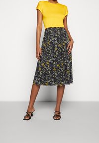Gelbes, tailliertes Oberteil kombiniert mit einem schwarzen, blumigen Midikleid, das gelbe und blaue Blumen zeigt. Das Model trägt schwarze Riemchensandalen.