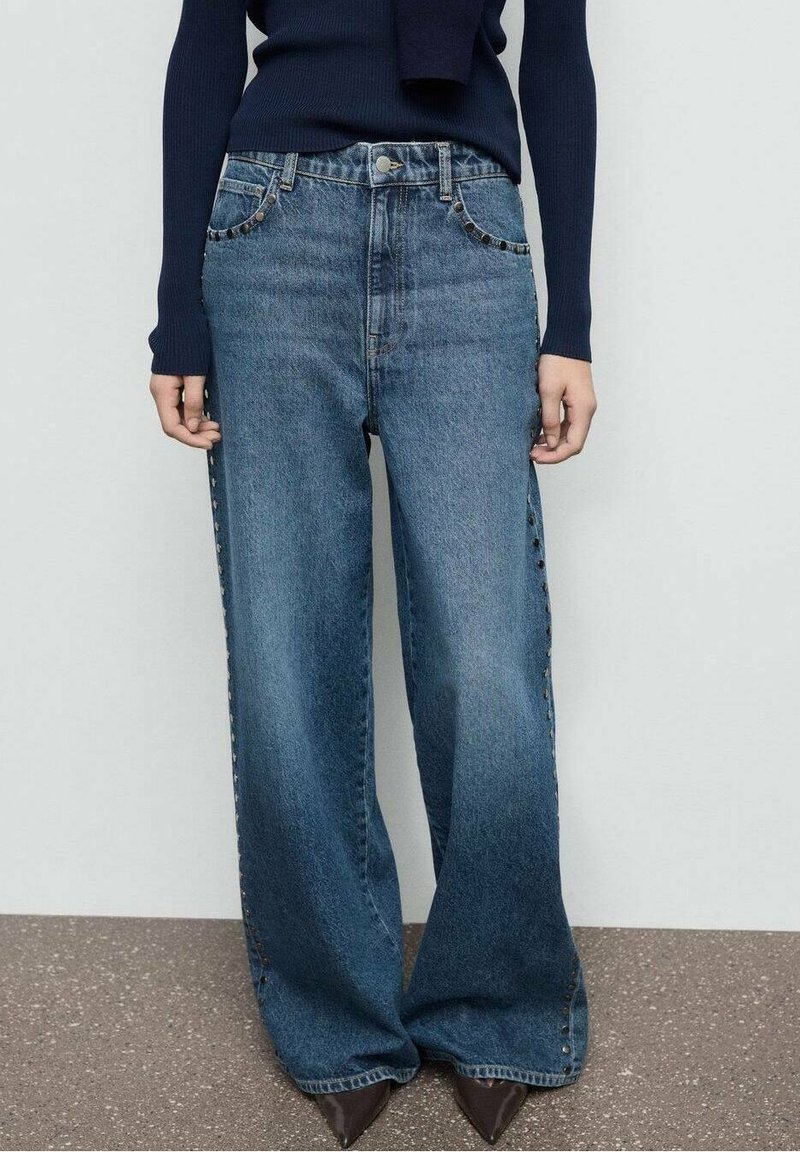 Personne portant un jean bleu taille haute à jambes larges avec des clous sur les côtés et les poches, associé à un haut ajusté bleu marine à manches longues et des chaussures pointues foncées.