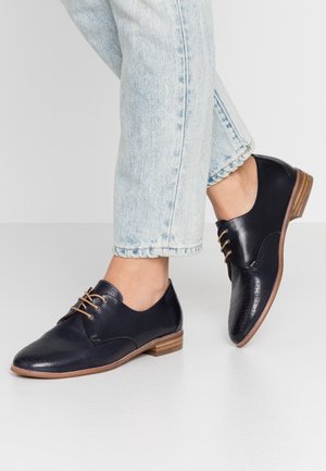 Sorte læder oxford-snøresko med perforerede detaljer båret med lyseblå cropped jeans på en ensfarvet lys baggrund.