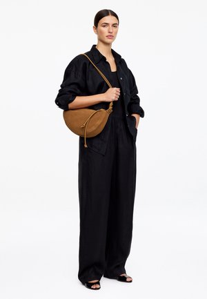 Vrouw draagt een losse zwarte outfit en zwarte sandalen, houdt een bruine suède schoudertas vast tegen een effen witte achtergrond.