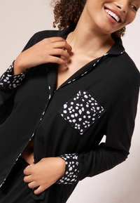 Černá pyžamová košile s límečkem, s kapsou na levé straně hrudi a vzorovanými rukávy v bílých a černých puntících.