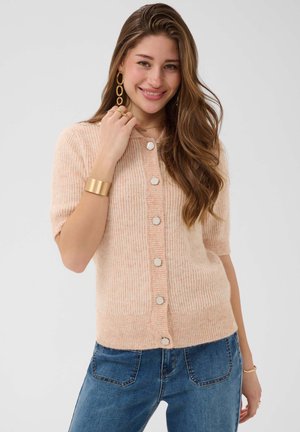 Jeune femme aux longs cheveux bruns portant un cardigan pêche à manches courtes boutonné et un jean bleu, souriante et touchant son visage.