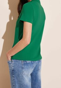 Grön pikétröja med korta ärmar, tillverkad av mjukt tyg, i kombination med ljusblå slitna denimjeans. Inga synliga mönster eller metallföremål.