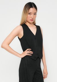 Gilet sans manches noir avec un décolleté en V profond, présentant un design ajusté et trois boutons sur le devant, associé à un pantalon à rayures.