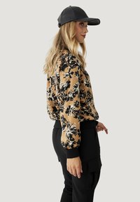 Blusa de manga larga floral en beige con patrones negros, combinada con pantalones cargo negros, que cuentan con puños acanalados y una gorra negra.