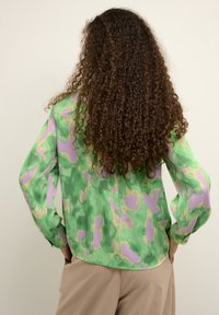 Persona con cabello largo y rizado, vistiendo una blusa de patrón verde y morado y pantalones beige, de pie con las manos en las caderas, de espaldas.
