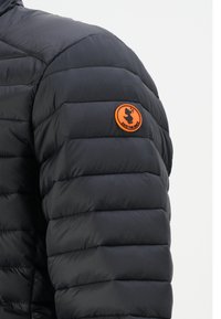 Zwarte pufferjack met horizontale stitching en een gestructureerd oppervlak. Heeft een ronde, oranje patch met een logo op de linkerschouder.