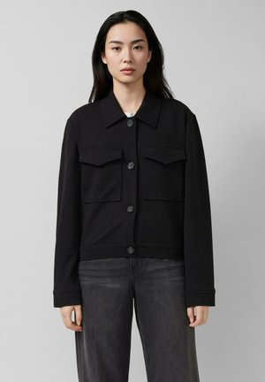 Jeune femme aux longs cheveux foncés portant une veste noire boutonnée avec des poches poitrine et un jean foncé, debout devant un fond gris.