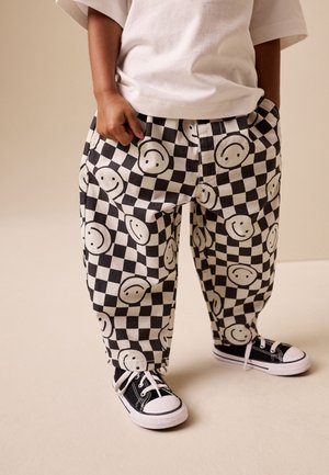 Enfant portant un pantalon à carreaux noir et blanc avec des visages souriants et des baskets en toile noires avec des lacets et semelles blanches.