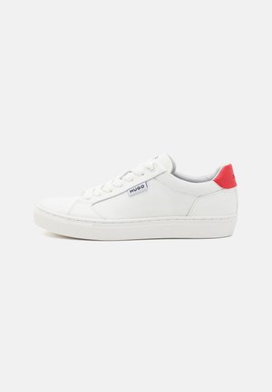 Sneaker bianca in materiale canvas, suola in gomma e accentuazione rossa contrastante sul tallone. Presenta otto occhielli e un sottile marchio laterale.