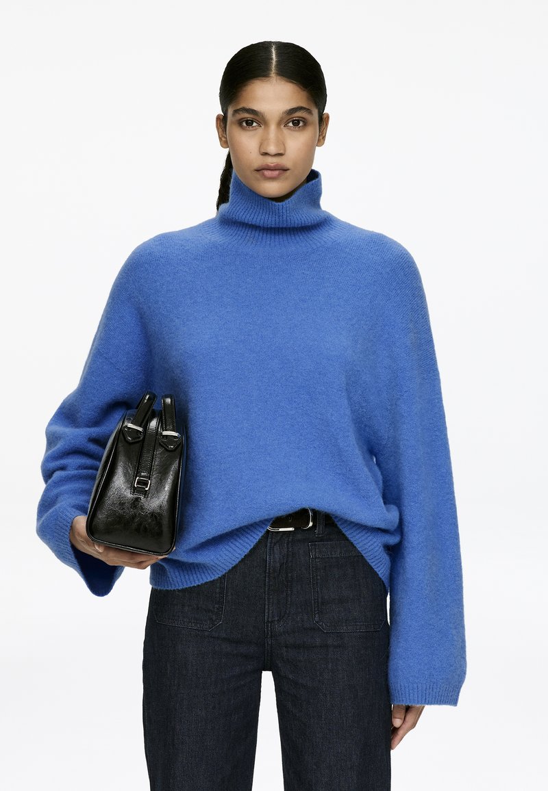 Oversized blue knit turtleneck sweater with long sleeves, paired with dark denim jeans and a small black leather bag with silver hardware.