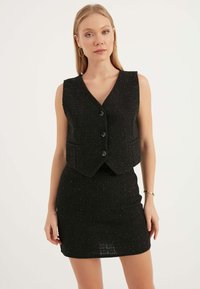 Svart tweedväst med V-ringning, fem knappar och matchande figursydd mini kjol. Texturerad tyg med subtila glansiga detaljer.