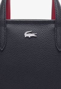 Sac en cuir noir texturé avec un intérieur rouge, orné d'un logo en crocodile argenté sur le devant. Les poignées sont robustes et arrondies.