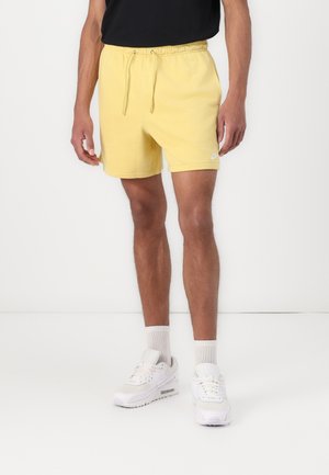 Mann trägt gelbe Shorts mit Kordelzug, weiße Socken und weiße Turnschuhe, steht vor einem einfarbigen weißen Hintergrund.