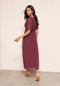 Bordová maxi šaty s krátkymi puff rukávmi, úzkym pásom a splývavou sukňou. Nosí sa s zlatými sandálmi na podpätku. Hladká, ľahká látka.