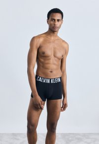 Μαύρες μποξερίτσες Calvin Klein με λευκή ζώνη logo, εφαρμοστό σχέδιο και μαλακό βαμβακερό ύφασμα, φορεμένες από μοντέλο χωρίς πουκάμισο.