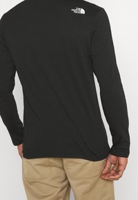 Černé tričko s dlouhými rukávy, kulatým výstřihem a bílým logem "The North Face" na horní části zad. Hladká látka a přiléhavý střih.